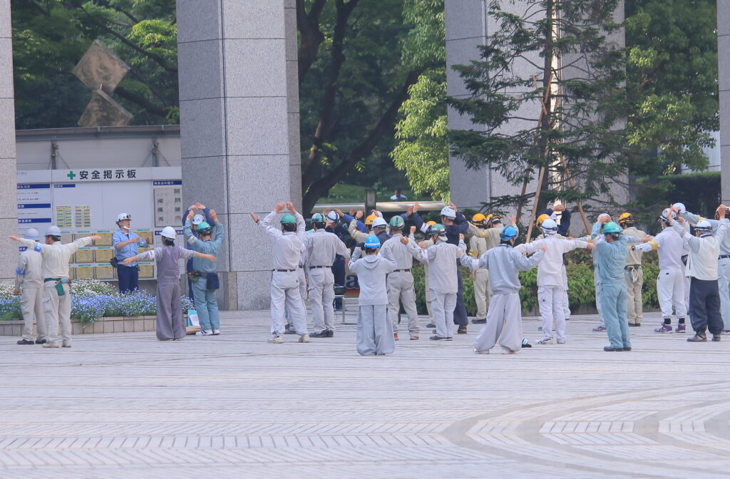 Groupe d’ouvriers en tenue de travail et casques de sécurité effectuant des exercices d’échauffement en extérieur avant de commencer leur journée.